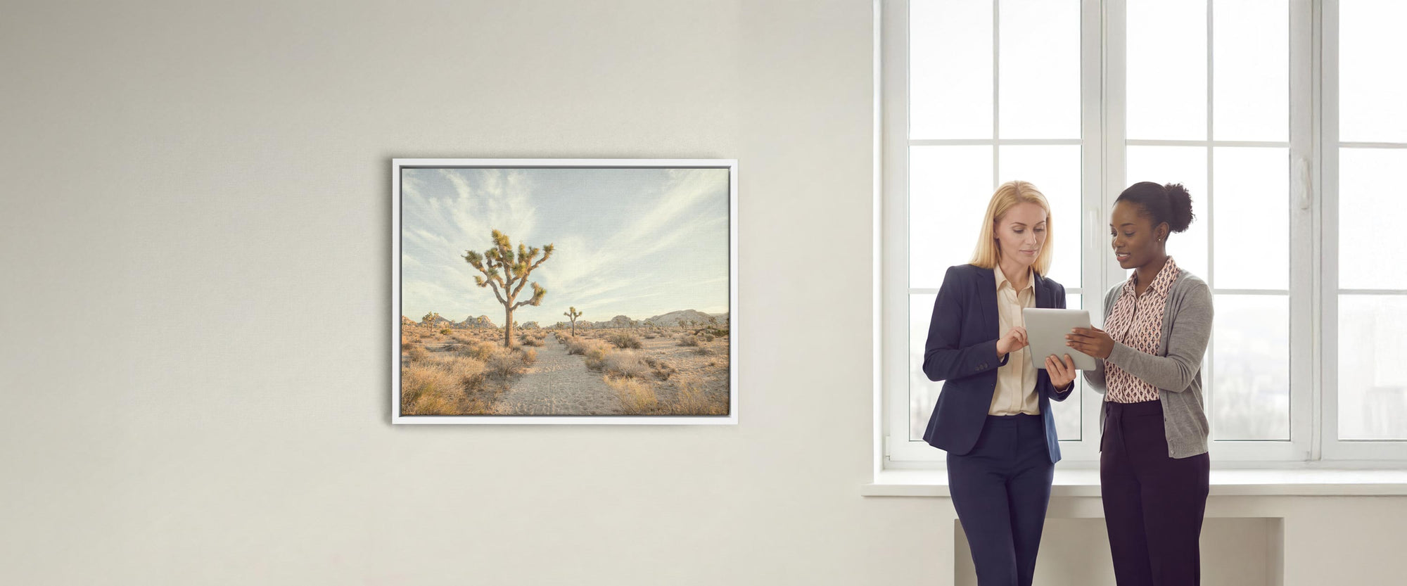 Two women standing in an office workspace with a large window, looking at a tablet next to a framed canvas wall art of a Joshua Tree landscape.