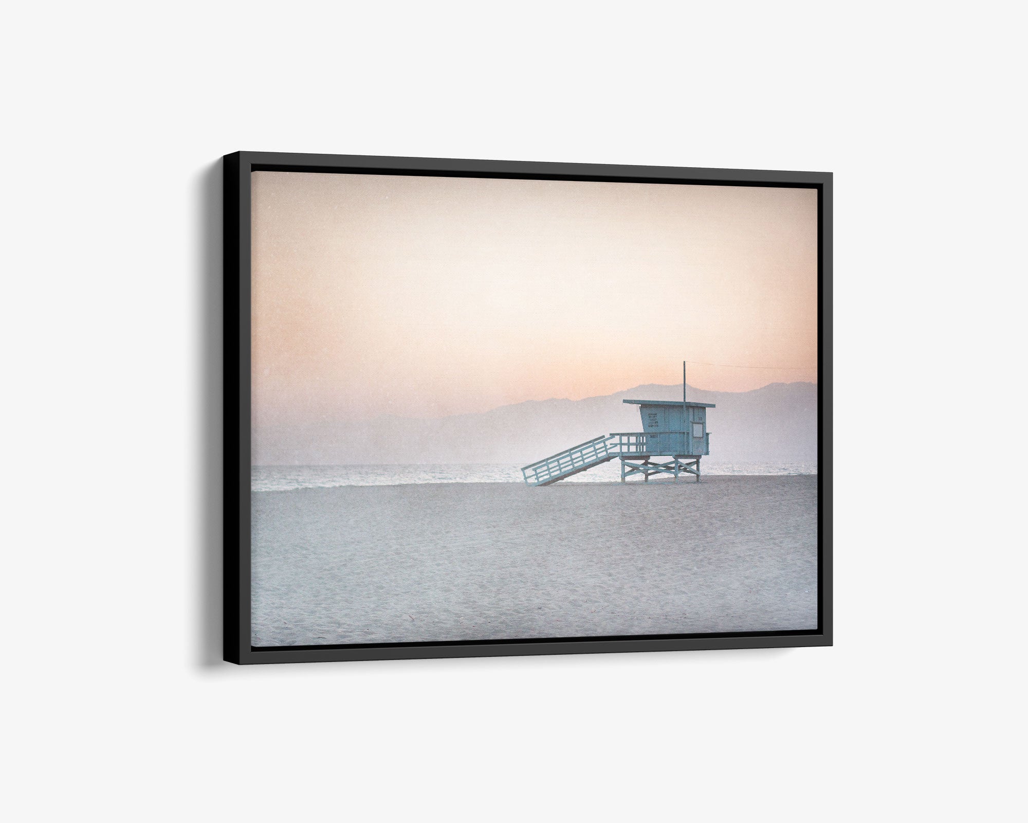 A framed sunset photograph depicting a lifeguard tower on serene Venice Santa Monica beach, with gentle hues of pink and blue in the sky and a faint mountain silhouette in the background - Offley Green&#39;s Pink Coastal Wall Art, &#39;Lifeguard Tower&#39;.