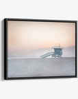 A framed sunset photograph depicting a lifeguard tower on serene Venice Santa Monica beach, with gentle hues of pink and blue in the sky and a faint mountain silhouette in the background - Offley Green's Pink Coastal Wall Art, 'Lifeguard Tower'.