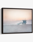 A framed Pink Coastal Wall Art of a lifeguard station at Venice Santa Monica Beach, with soft pastel hues in the sky and serene water, mounted on a white wall by Offley Green.