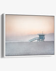 A framed Pink Coastal Wall Art photograph of a 'Lifeguard Tower' on Venice Santa Monica Beach by Offley Green, with gentle hues of pink and blue in the sky and a soft glow reflecting off the ocean.