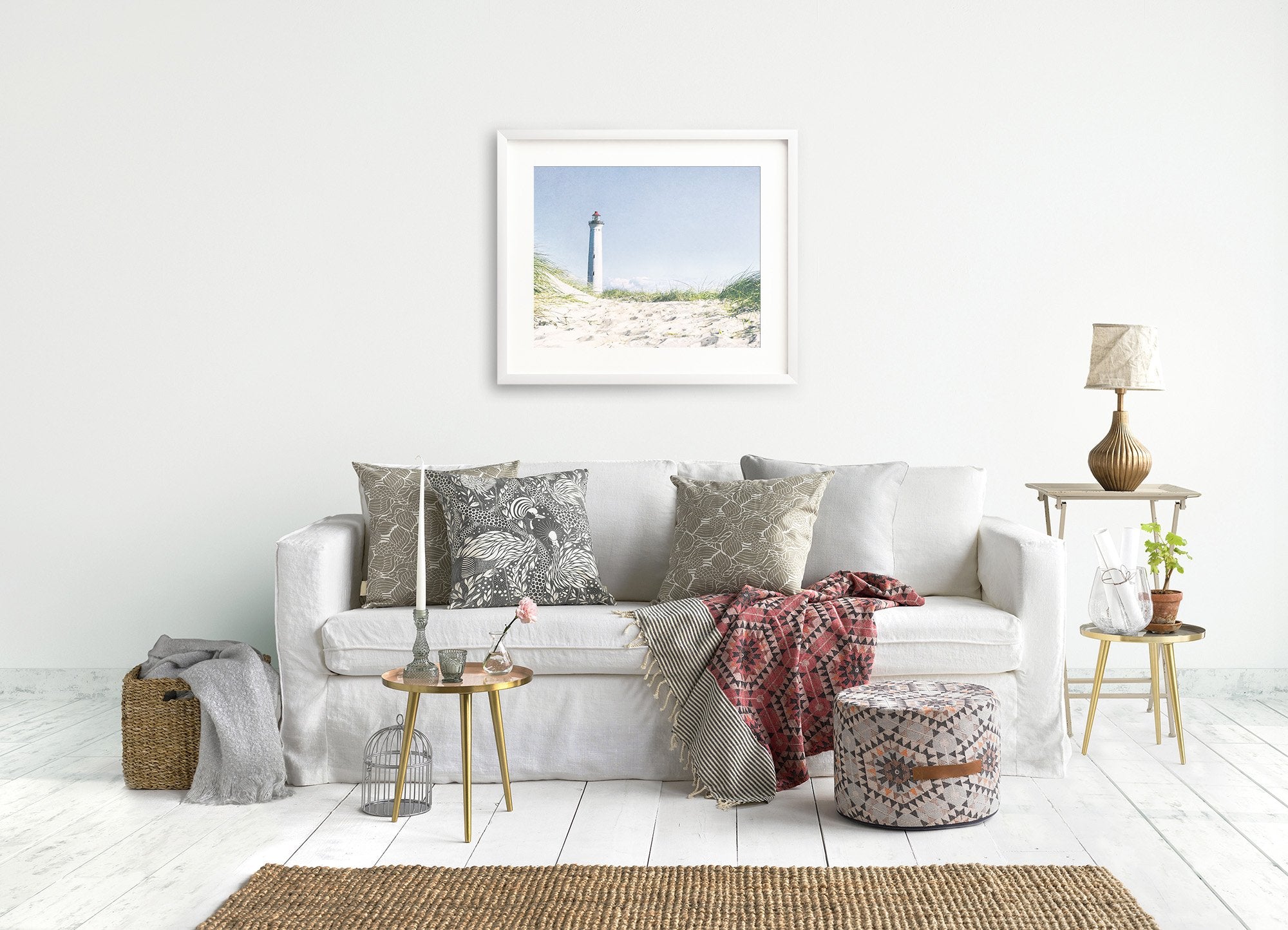 A serene living room featuring a white sofa adorned with various patterned cushions, a small round table, a wooden floor lamp, Offley Green's Nautical Print, 'The Lighthouse' on the wall, and decorative items including plants and a woven basket.