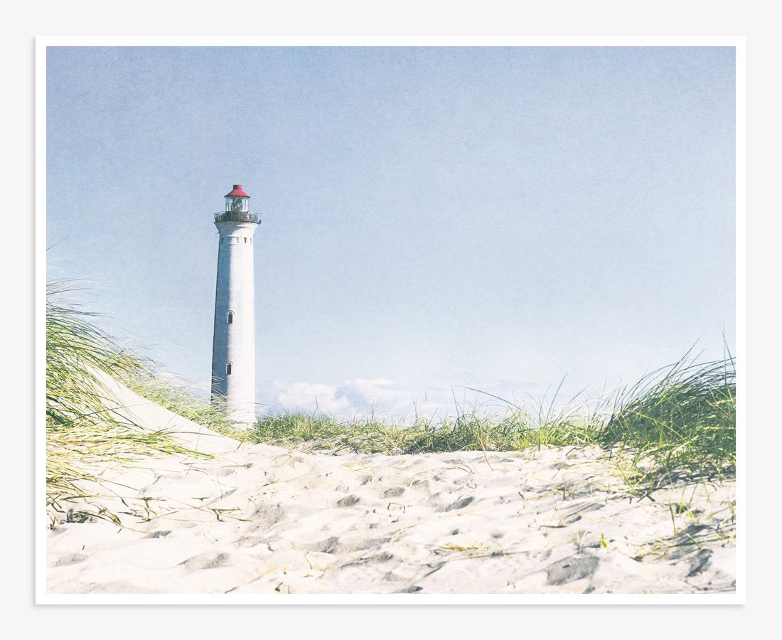 A picturesque white lighthouse stands tall against a clear sky, viewed from a sandy beach with tufts of green grass in the foreground, perfect for coastal decor with Offley Green&#39;s Nautical Print, &#39;The Lighthouse&#39;.