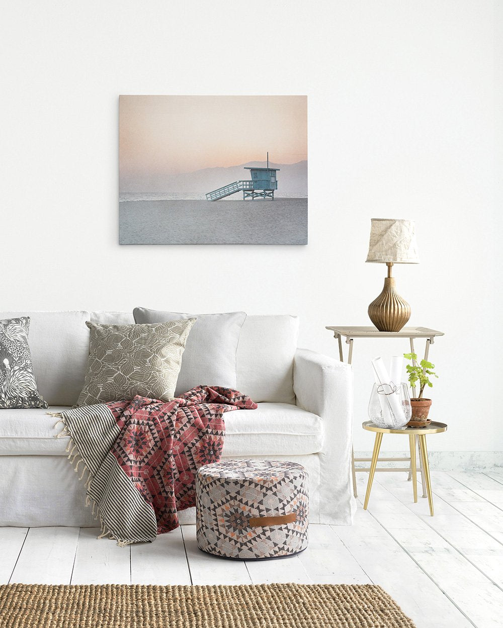 A cozy living room featuring a white sofa with decorative pillows, a woven pouf, a side table with a lamp and plant, and Offley Green&#39;s Pink Coastal Wall Art, &#39;Lifeguard Tower&#39; print of Venice Santa Monica beach at sunset on the wall.