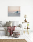A cozy living room featuring a white sofa with decorative pillows, a woven pouf, a side table with a lamp and plant, and Offley Green's Pink Coastal Wall Art, 'Lifeguard Tower' print of Venice Santa Monica beach at sunset on the wall.