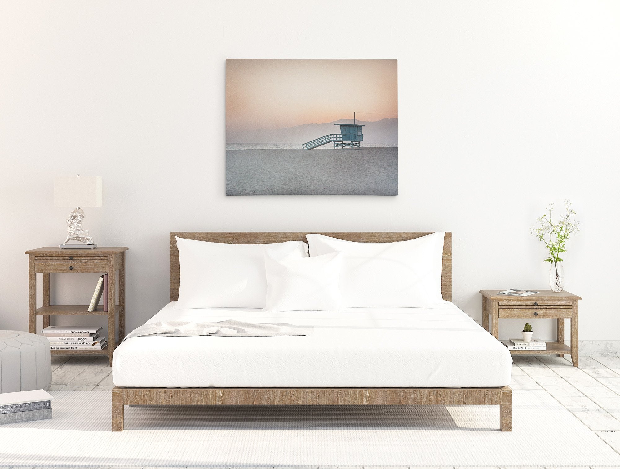 A minimalist bedroom featuring a neatly made bed with white bedding, flanked by wooden nightstands with lamps and a plant. A framed Sunset photograph of Venice Santa Monica beach hangs above the bed from Offley Green&#39;s Pink Coastal Wall Art, &#39;Lifeguard Tower&#39;.