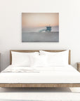 A minimalist bedroom featuring a neatly made bed with white bedding, flanked by wooden nightstands with lamps and a plant. A framed Sunset photograph of Venice Santa Monica beach hangs above the bed from Offley Green's Pink Coastal Wall Art, 'Lifeguard Tower'.