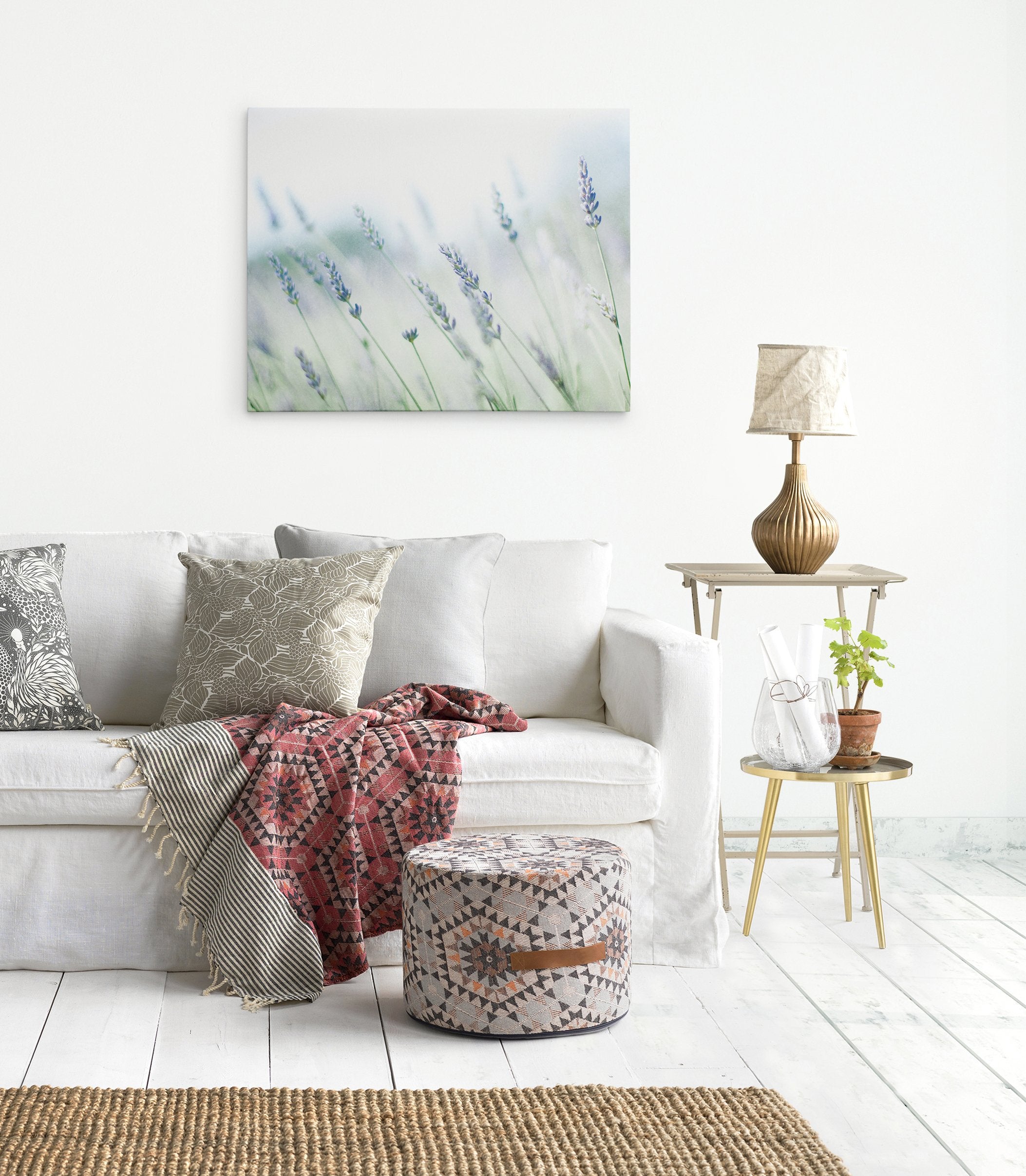 A cozy living room corner with a white sofa adorned with colorful cushions, a knit blanket, a small wood side table with a lamp, and a large Offley Green canvas gallery wrap of Rustic Farmhouse Canvas Wall Art, &#39;Buds of Lavender&#39; on the wall.