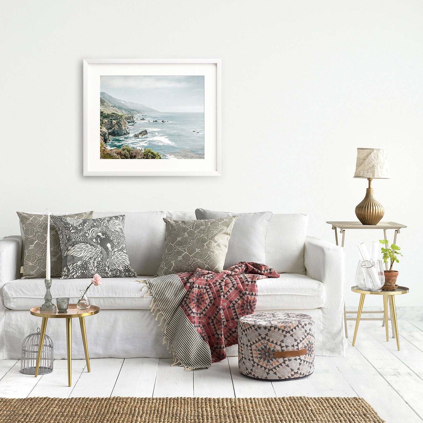 A cozy living room with a white sofa decorated with patterned cushions, a side table with a lamp, a basket, and an Offley Green Big Sur Landscape Print of &#39;Rocky Rocks&#39; on the wall.