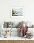 A cozy living room with a white sofa decorated with patterned cushions, a side table with a lamp, a basket, and an Offley Green Big Sur Landscape Print of 'Rocky Rocks' on the wall.