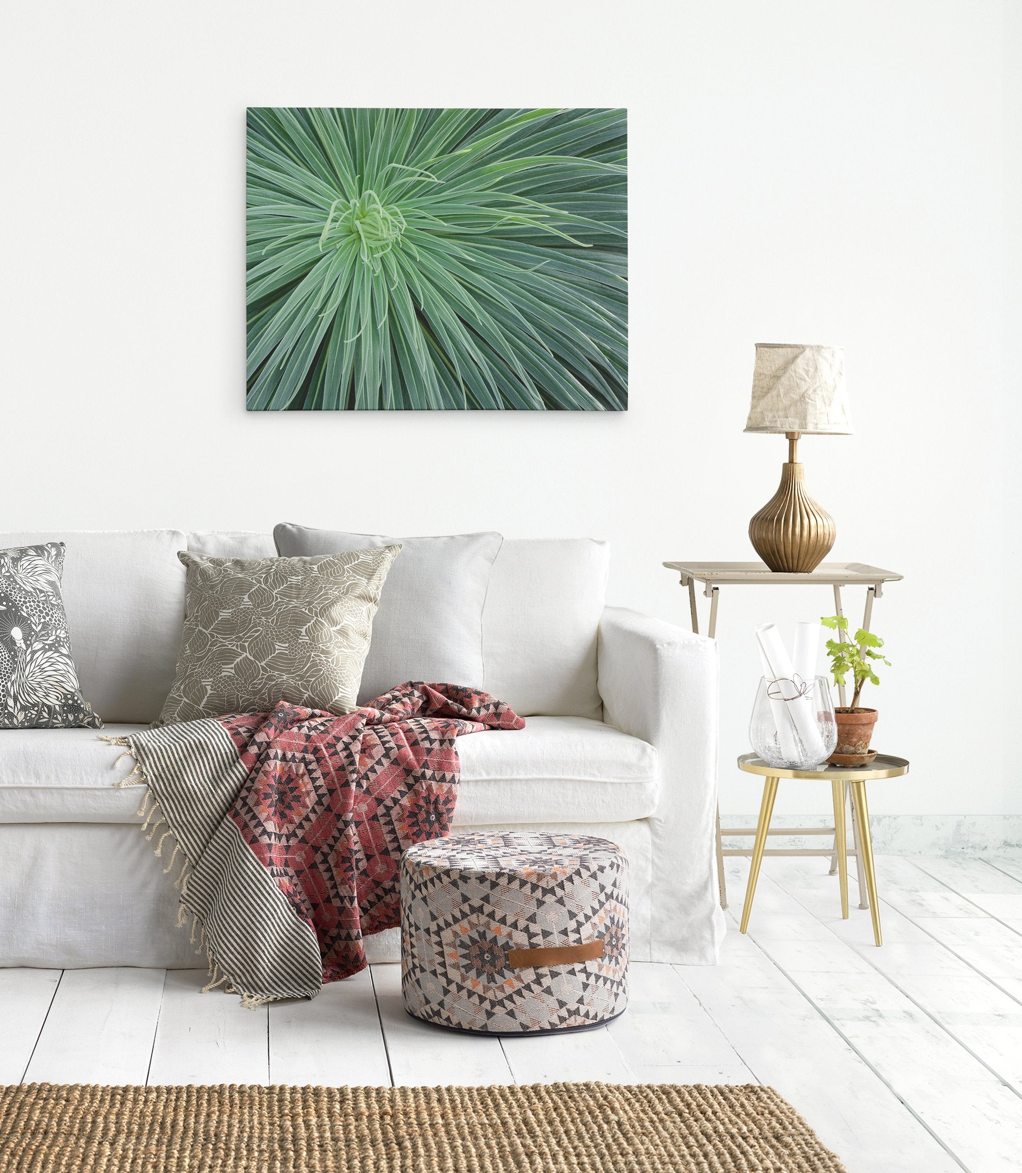 A cozy living room corner featuring a white sofa with decorative pillows, a patterned ottoman, a wooden side table with a lamp, and an Offley Green Abstract Green Botanical Canvas Wall Art, 'Desert Fireworks' with desert plants image