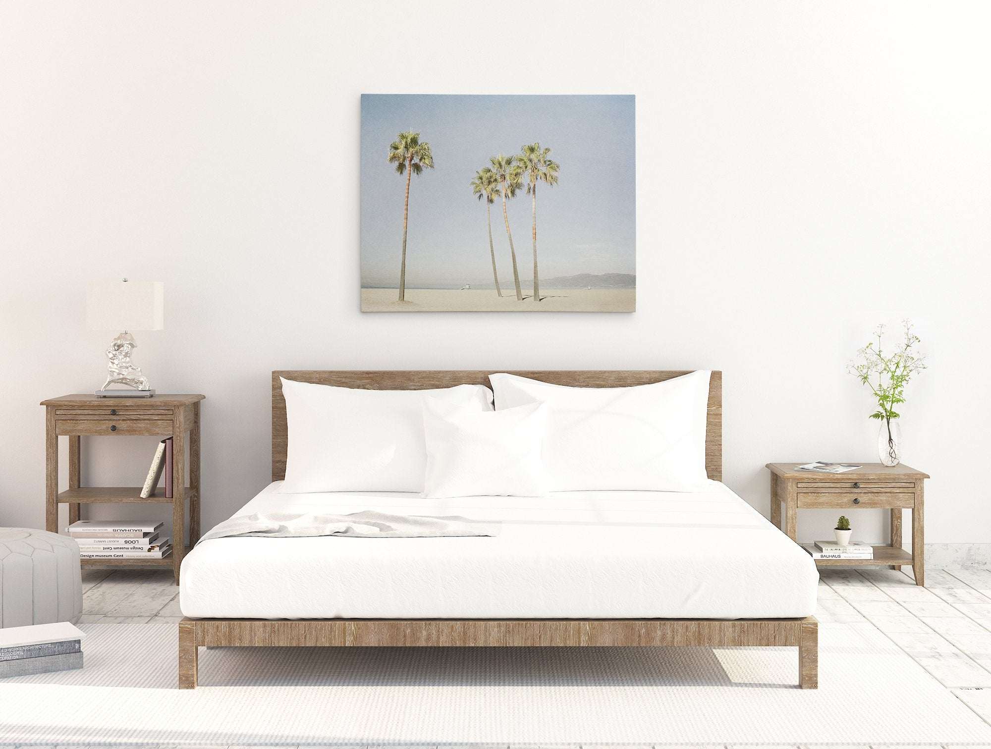 Modern bedroom with a minimalist design featuring a wooden bed frame with white bedding. Flanked by two wooden nightstands, one with a lamp and the other with a vase of flowers. Above the bed hangs an Offley Green California Venice Beach Canvas Wall Art, &#39;Boardwalk Palms&#39;, rendered in exquisite detail.