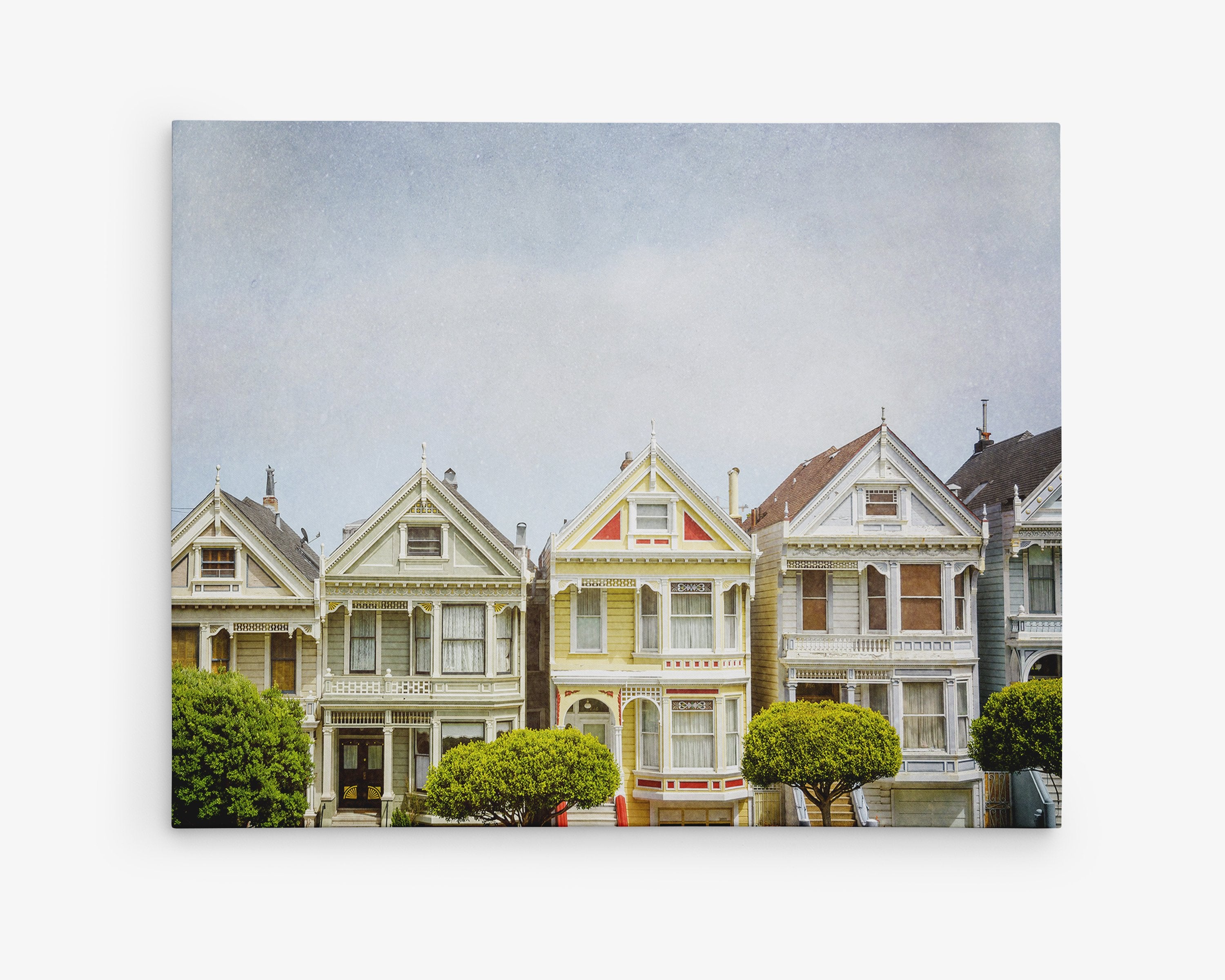 A row of five colorful Victorian houses, known as the Painted Ladies, stands closely together in San Francisco. Each house has distinct ornate details and pastel colors ranging from yellow and blue to red and beige. Two trimmed bushes are in front of the houses under a cloudy sky, like Offley Green&#39;s San Francisco Canvas Wall Art, &#39;Painted Ladies&#39; come to life.