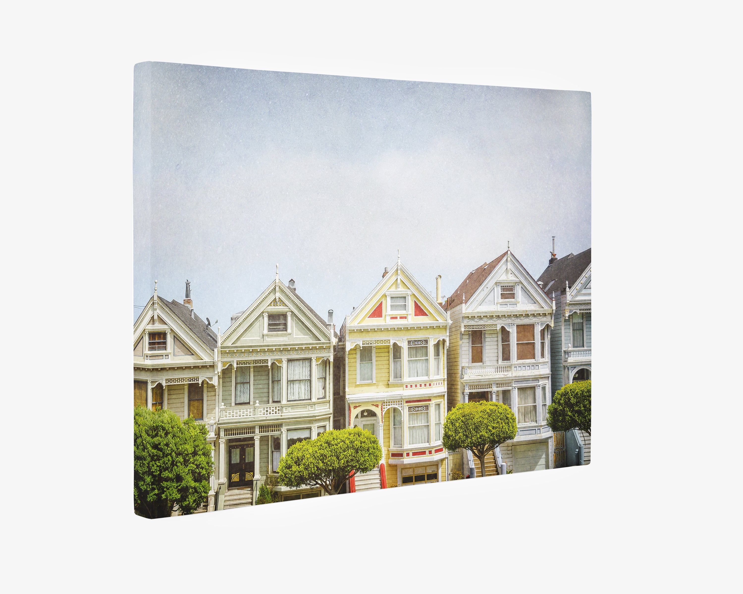 The San Francisco Canvas Wall Art, &#39;Painted Ladies&#39; by Offley Green depicts a row of colorful Victorian houses, known as the Painted Ladies in San Francisco, with intricate architectural details. Each house features ornate trim, bay windows, and gabled roofs. In the foreground, neatly trimmed trees add greenery to the scene. The sky is clear and light blue.