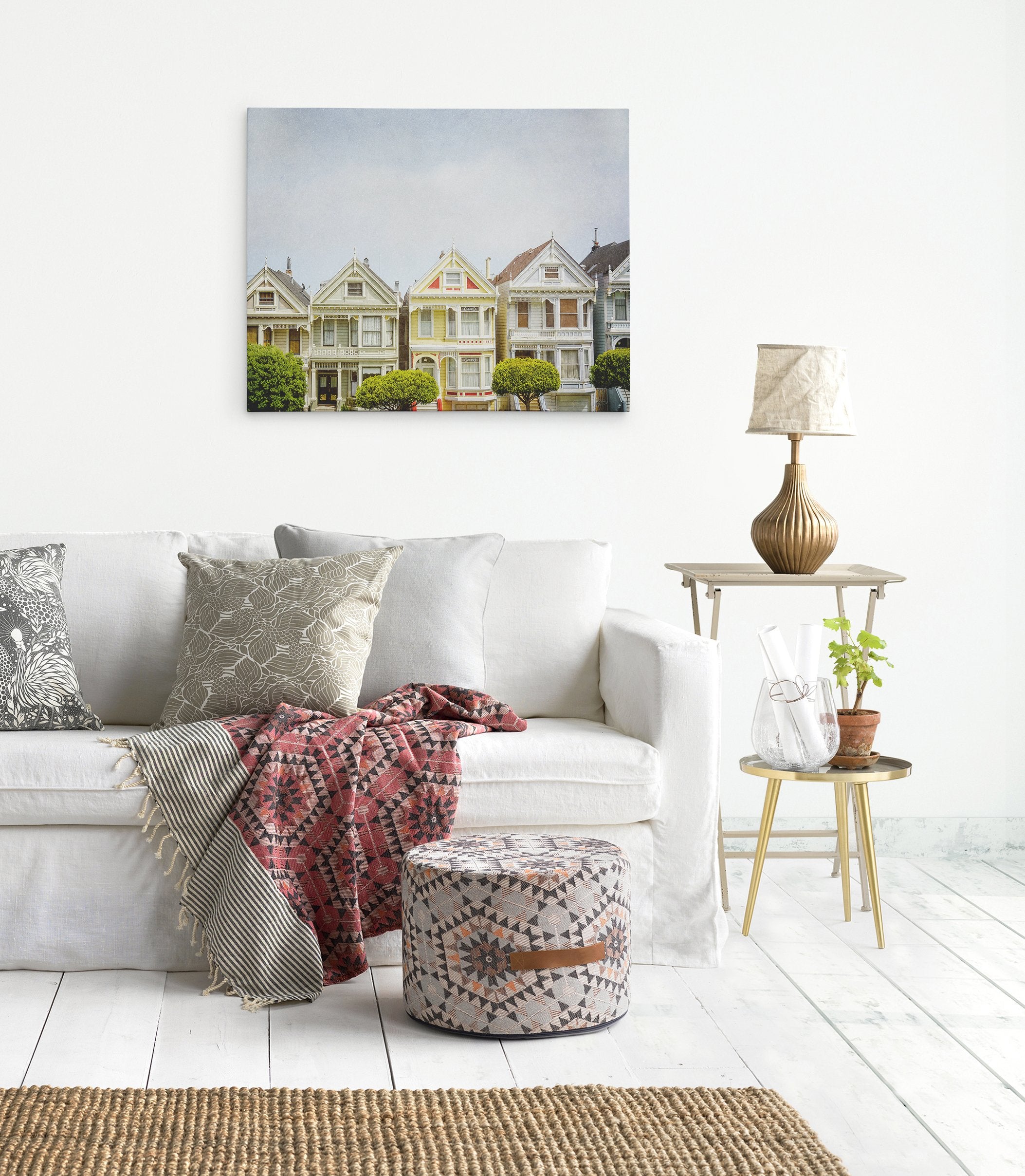 A cozy living room with a white sofa adorned with patterned throw pillows and a colorful blanket. A small table on the right holds a lamp and a plant. Above the sofa, an Offley Green San Francisco Canvas Wall Art, &#39;Painted Ladies&#39;, adds charm. A woven rug and a patterned pouf complete the space.