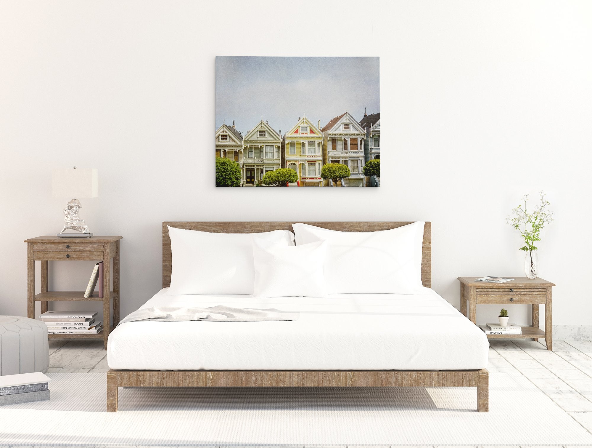 A minimalistic bedroom with a wooden bed frame and white bedding. Two matching wooden nightstands with lamps and decorative items flank the bed. A San Francisco Canvas Wall Art, &#39;Painted Ladies&#39; by Offley Green hangs on the white wall above the bed. The floor is light-colored wood.